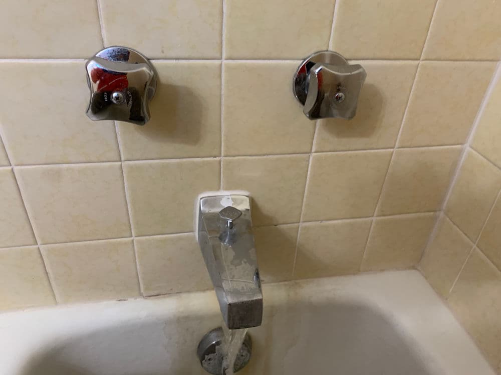 Chrome bathtub faucet with two separate knobs and running water against tiled wall.
